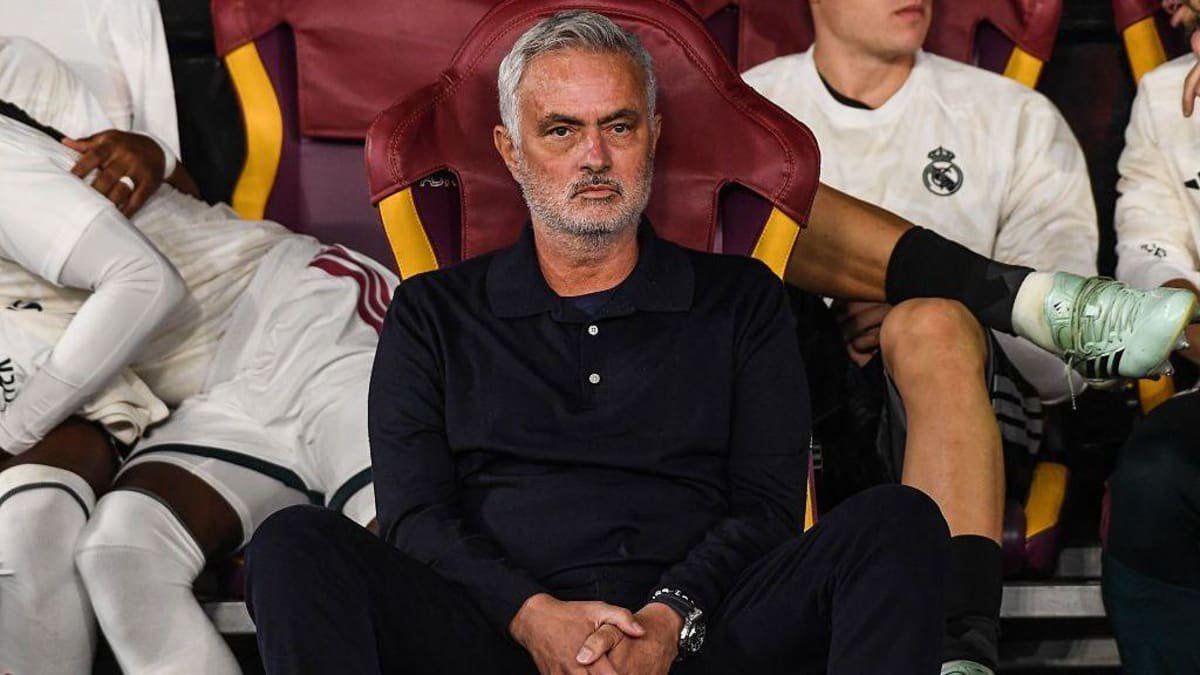 Jose Mourinho sits, straight-faced, on a seat in dark clothing, with Real Madrid players in white kit sitting behind him