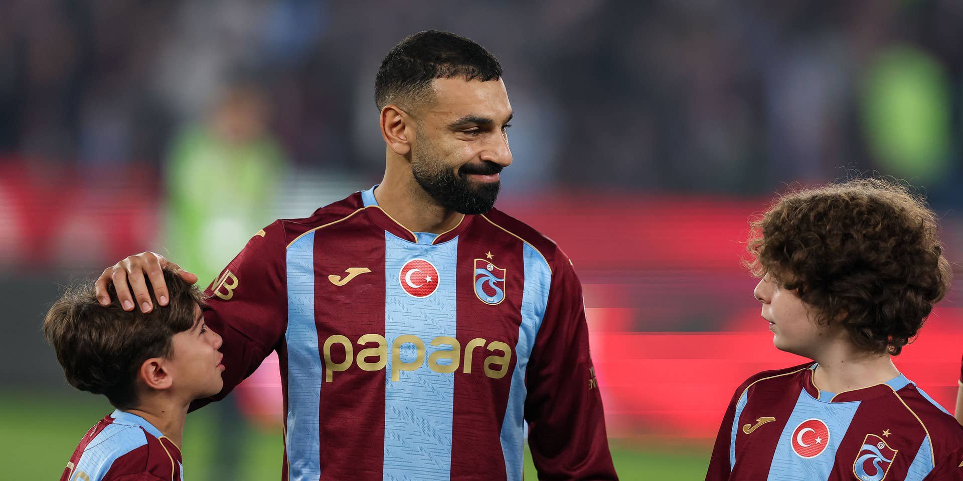 Mohamed Salah lächelt im rot-blau gestreiften Trikot von Trabzonspor auf dem Spielfeld und legt seine Hand sanft auf den Kopf eines jungen Einlaufkindes.
