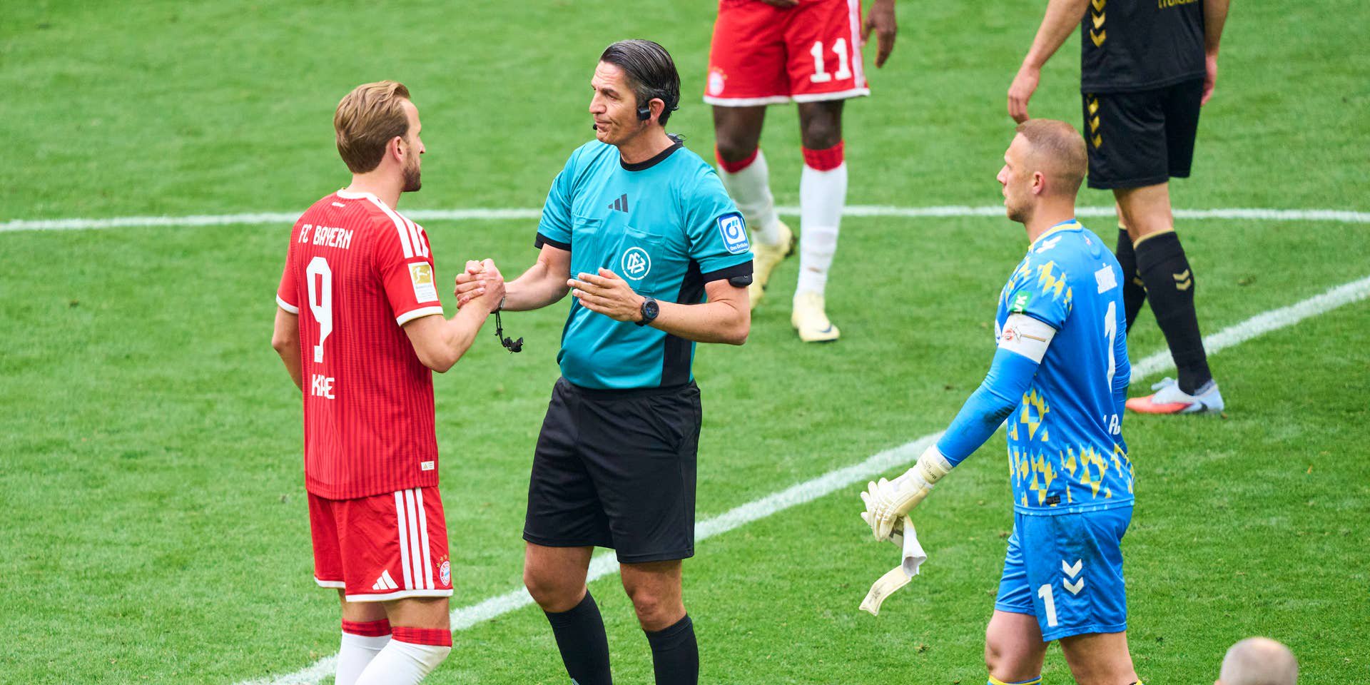 Schiedsrichter begrüßt einen Bayern-Spieler und einen Torwart nach Abpfiff auf dem Fußballfeld.