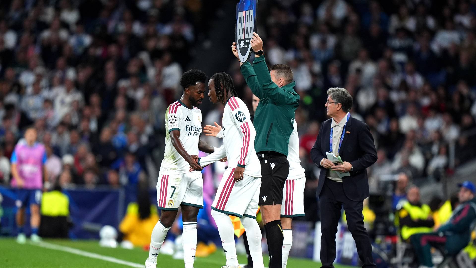 Dois jogadores de futebol, um com a camisa 7 e outro com dreadlocks, se cumprimentam no campo enquanto um árbitro levanta a placa de substituição e um homem de terno observa.