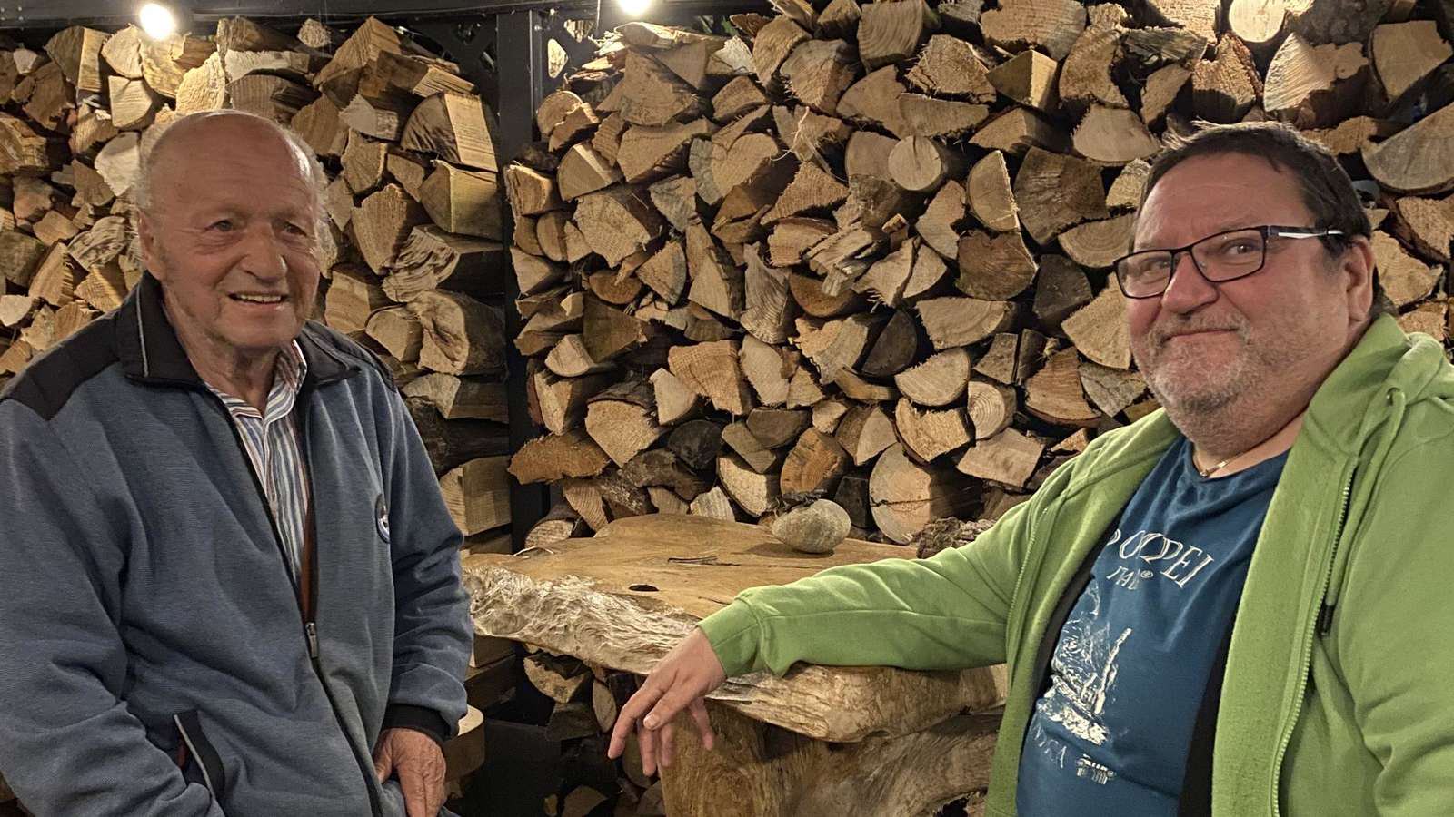 Skatmeister Jakob Jakel und Skatclubchef Mirko Berger vom Haager Skatclub Pik 7 sitzen an rustikalen Holzmöbeln vor aufgestapeltem Brennholz.