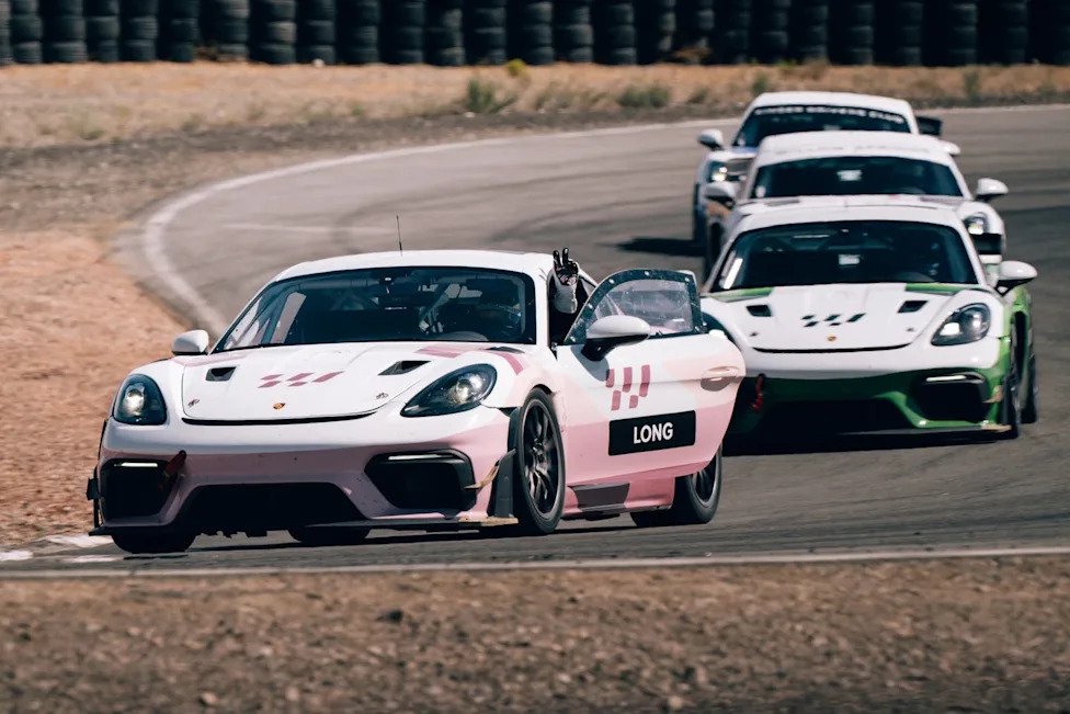 Race cars on a track, featuring a pink Porsche in the foreground with an open door and a raised hand from the driver.