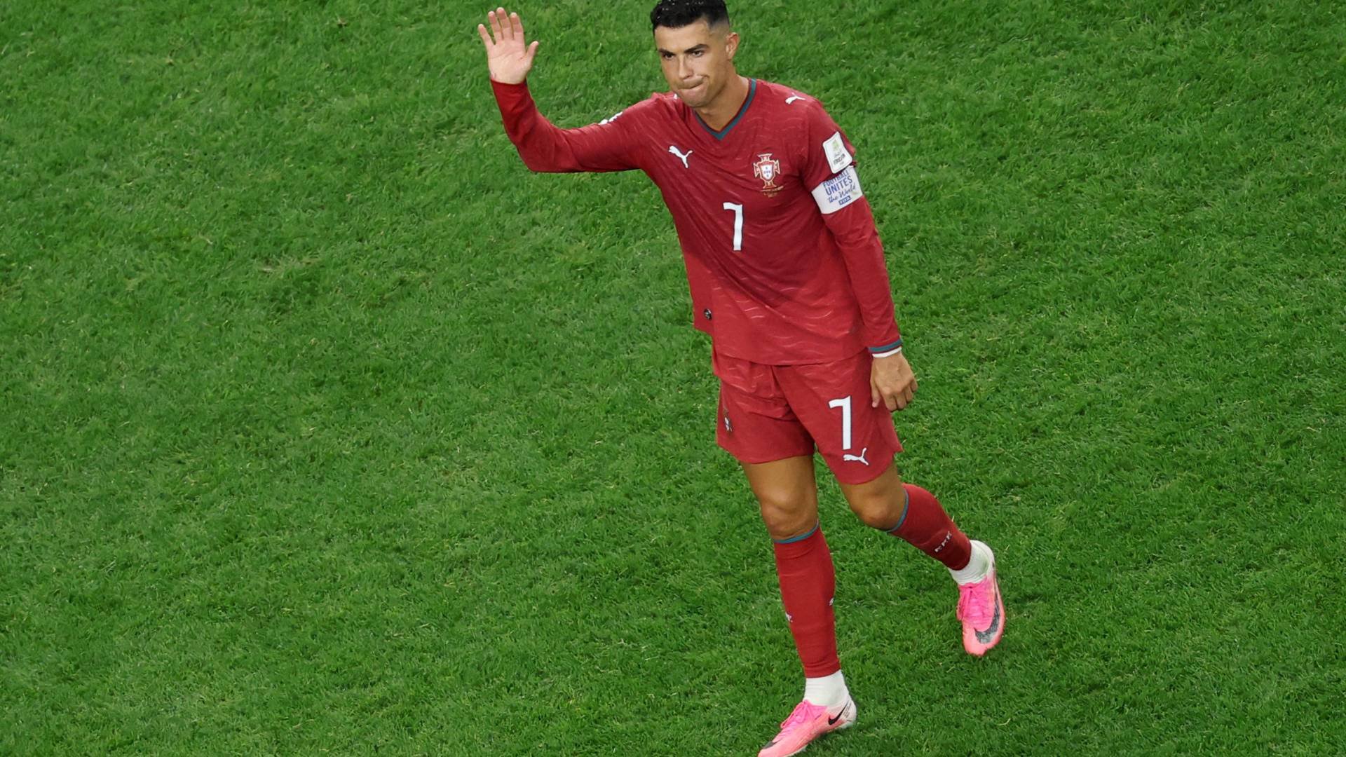 Cristiano Ronaldo, jogador de futebol, acena com a mão direita enquanto caminha sobre a grama verde do campo. Ele veste um uniforme vermelho com o número 7 e chuteiras rosa