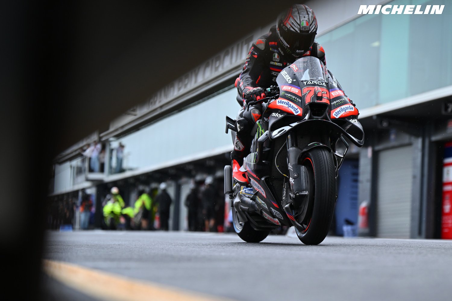 Aprilia MotoGP rider in black and red leathers on a Aprilia RS-GP, race number 72, riding down the pit lane