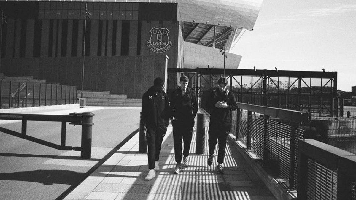 Three footballers, who are all wearing Everton tracksuits, are walking towards the camera at the Everton stadium in this candid black and white image. 