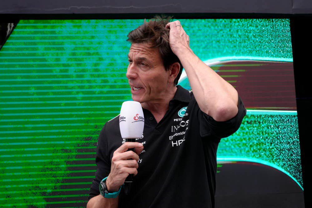 A man in a black Mercedes-AMG Petronas F1 team polo holds a microphone and scratches his head during a paddock interview at a Formula 1 event.