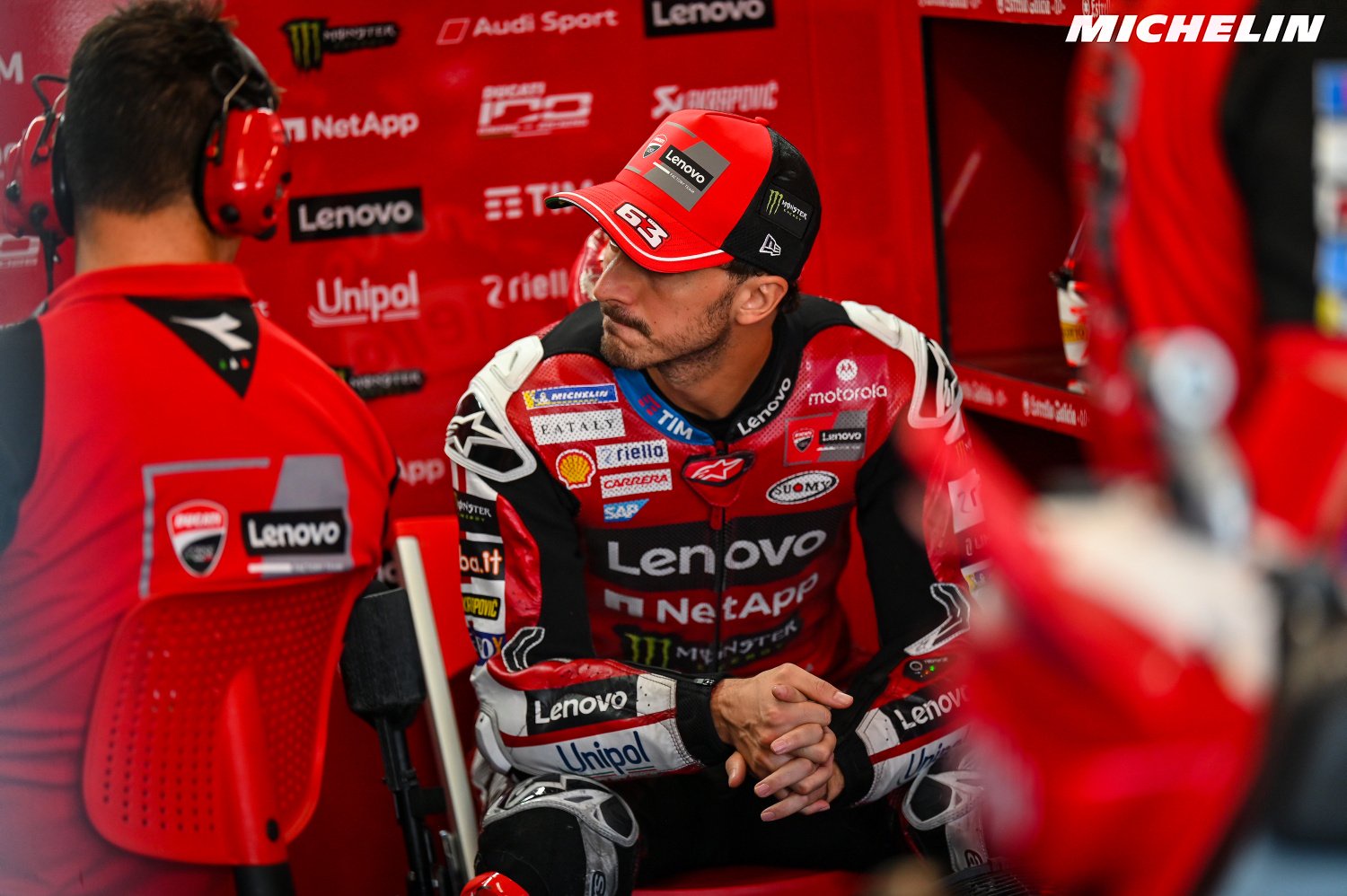 A Ducati MotoGP rider in red Lenovo and NetApp leathers and a red cap sits in the pit box, looking pensive.