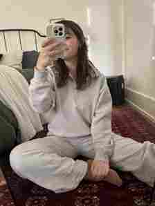 A person sitting on a red patterned rug, wearing a light gray tracksuit, holds up a phone to take a mirror selfie in a bedroom with a bed and white walls.