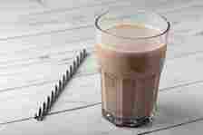 A glass of chocolate milk sits on a wooden surface next to a black-and-white striped straw