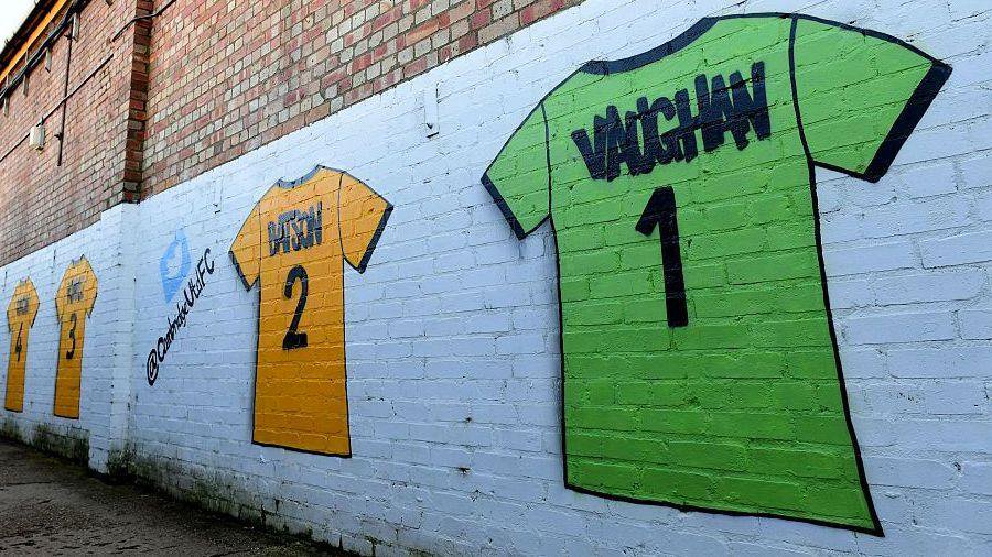 A mural on a brick wall shows amber Cambridge shirts and a green goalkeeper shirt with the name VAUGHAN and number one.