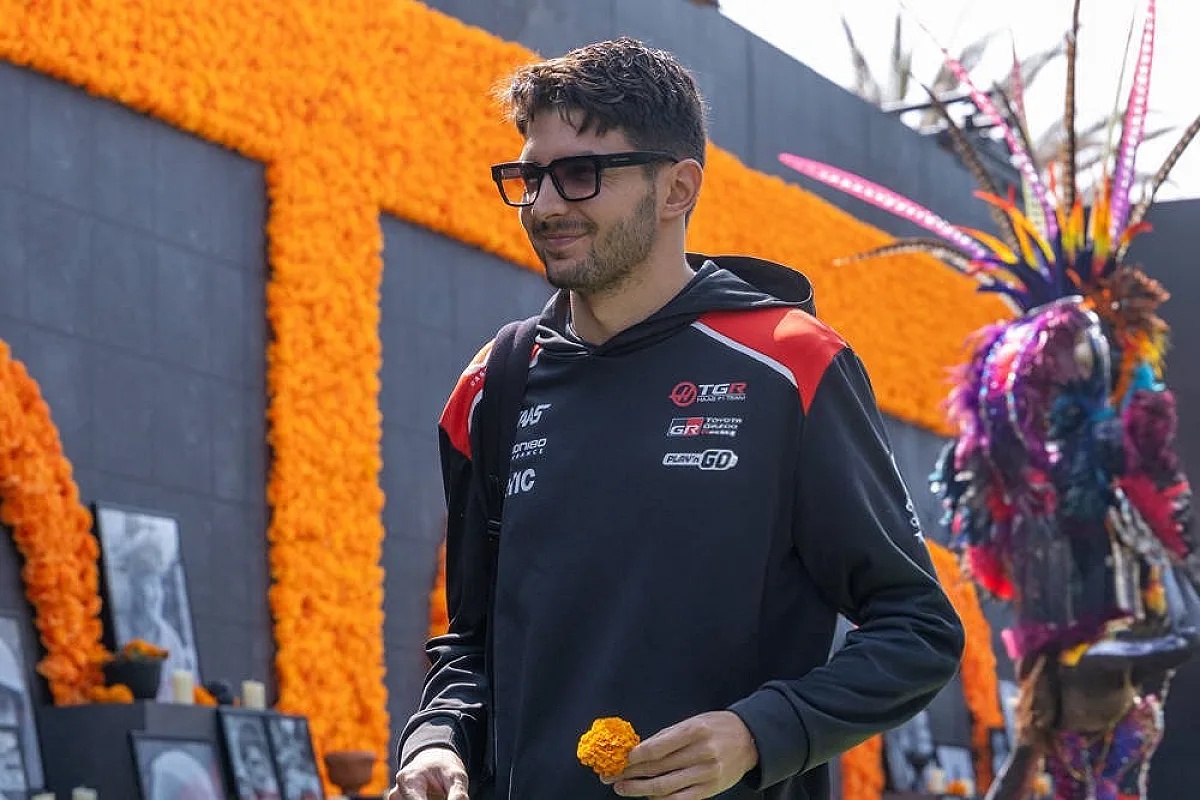 A driver in a black Toyota Gazoo Racing hoodie and dark glasses holds a marigold near colorful Day of the Dead ofrendas in a paddock setting.