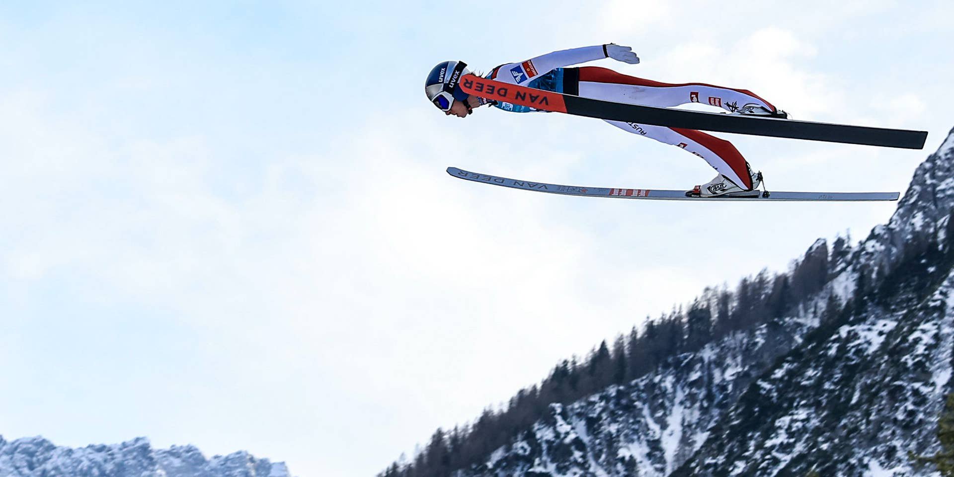 Ein Skispringer fliegt durch die Luft vor verschneiten Bergen und blauem Himmel.