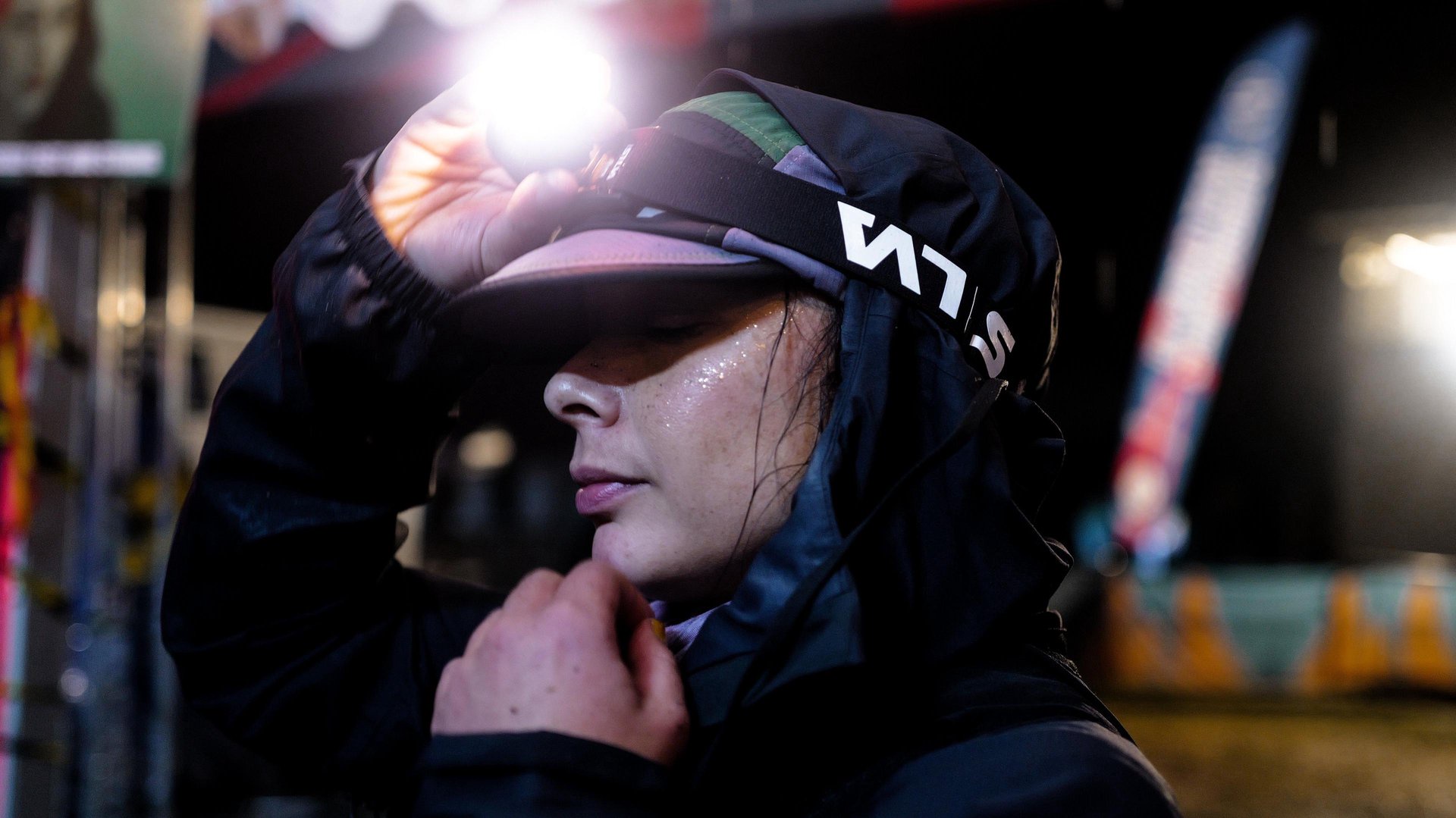 A close up of Sarah who is standing in a lit area at night time, facing side on. She is wearing a hat with a head torch which she is adjusting with her hand, and her hood is up over her head. Her other hand is touching her chin
