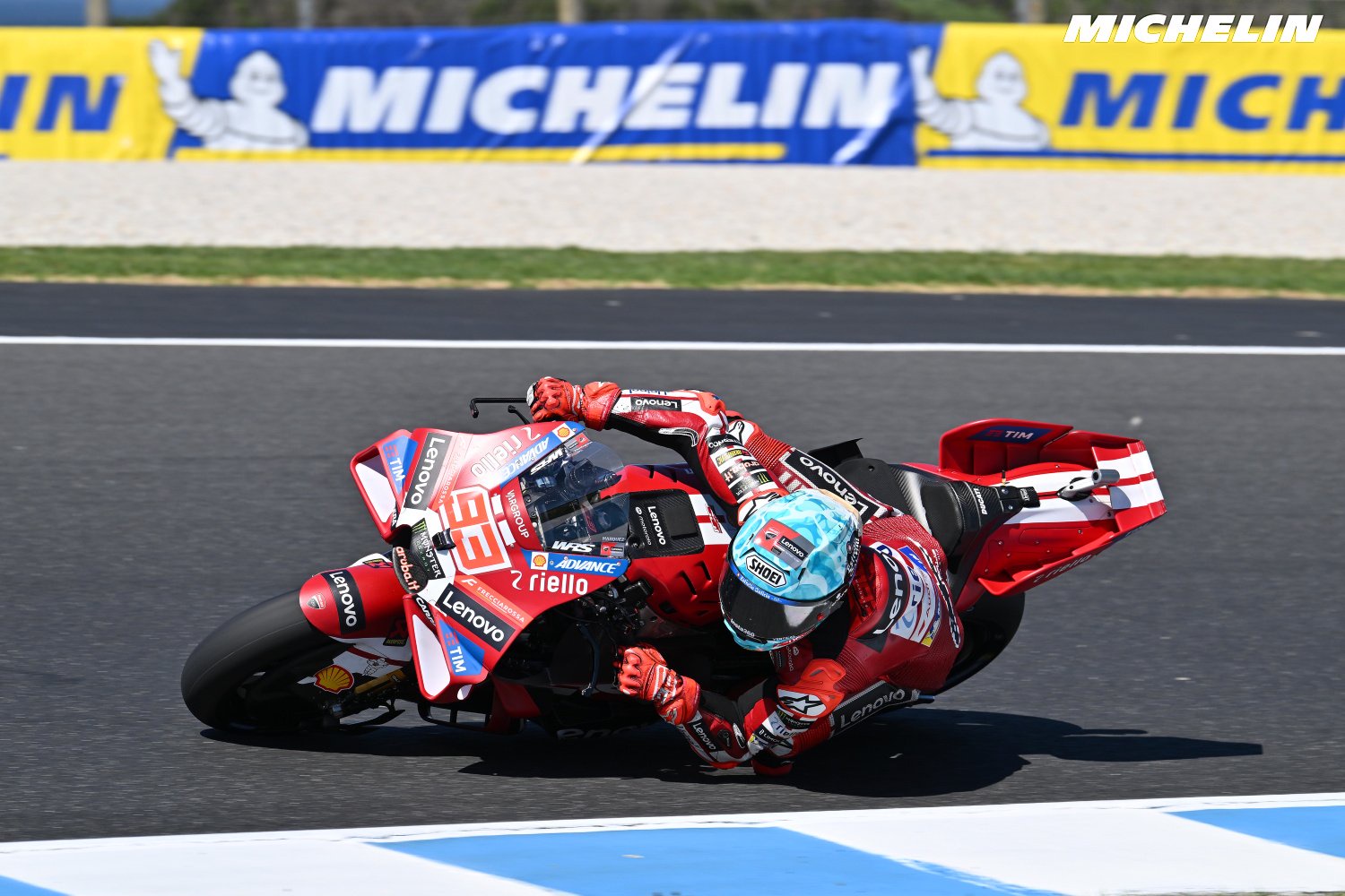 A rider in red Ducati leathers leans deep into a corner on a red Ducati MotoGP bike at Phillip Island, with Michelin barriers behind.