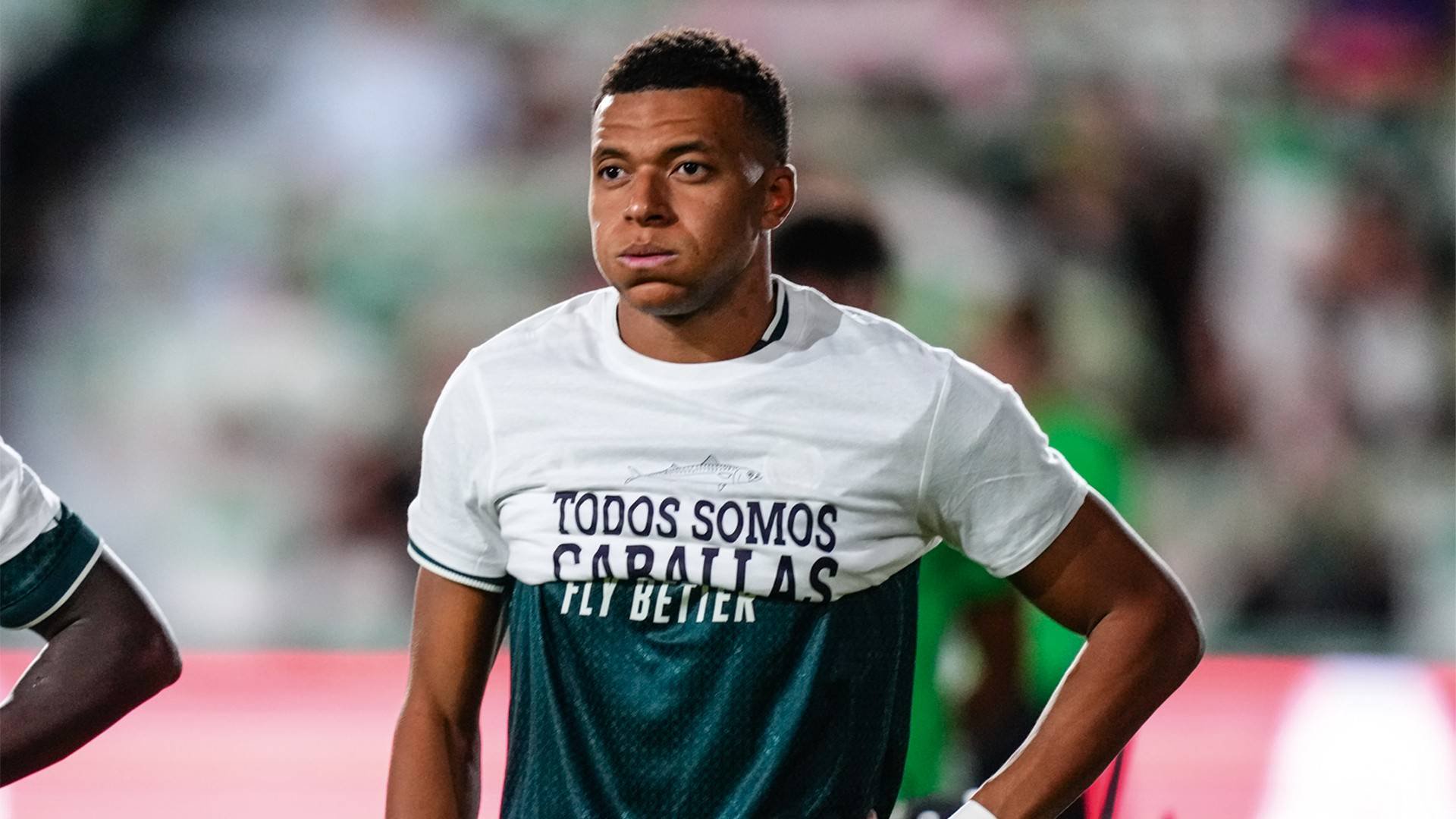 Jogador de futebol Kylian Mbappé, com expressão séria, vestindo uma camisa branca com a frase TODOS SOMOS CARALLAS FLY BETTER e uma camisa verde escura por baixo, em um campo de futebol com fundo desfocado