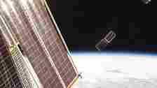  Two small, rectangular satellites deploy from the international space station with earth in the background. 