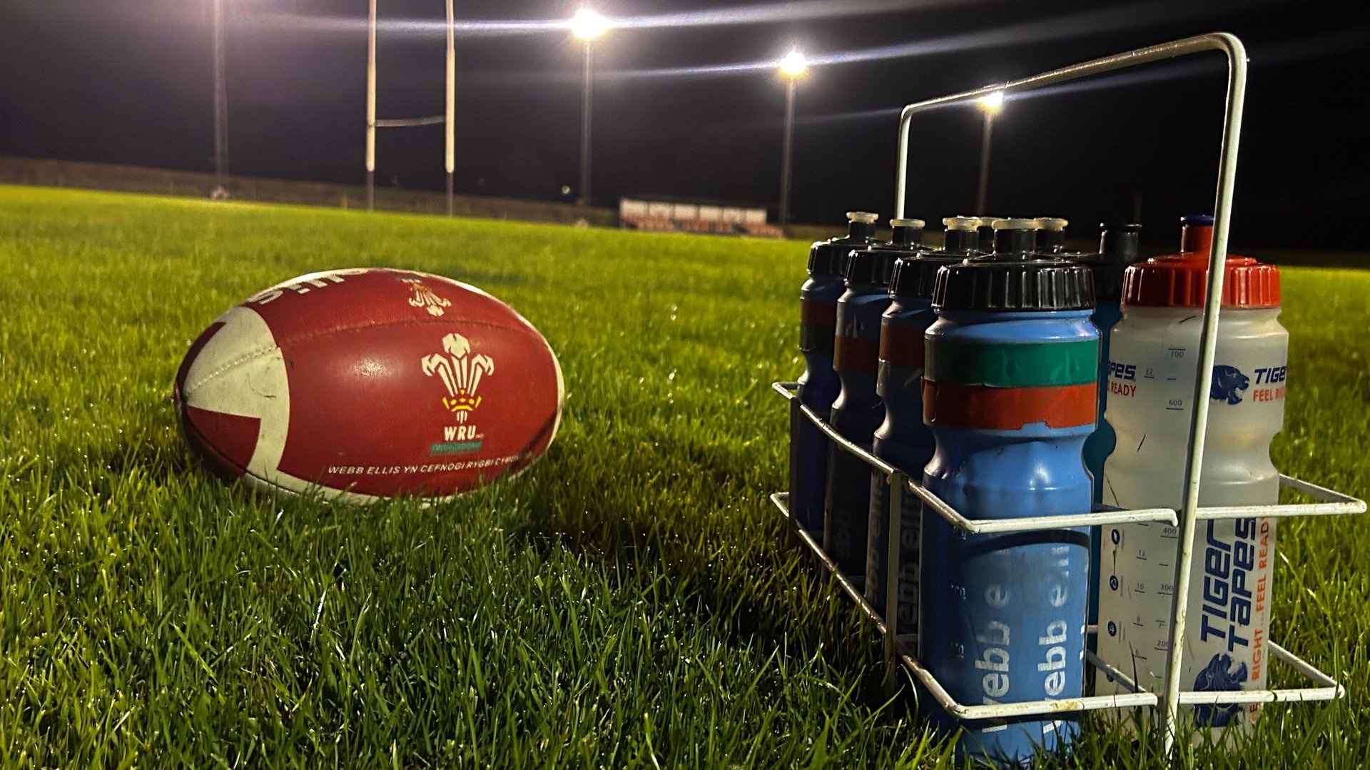 A rugby ball on a grassy rugby pitch, alongside several water bottles.