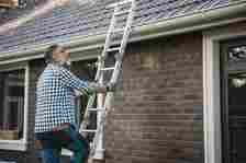 An older man in a plaid shirt and gloves climbs a ladder to clean the gutters on a brick house