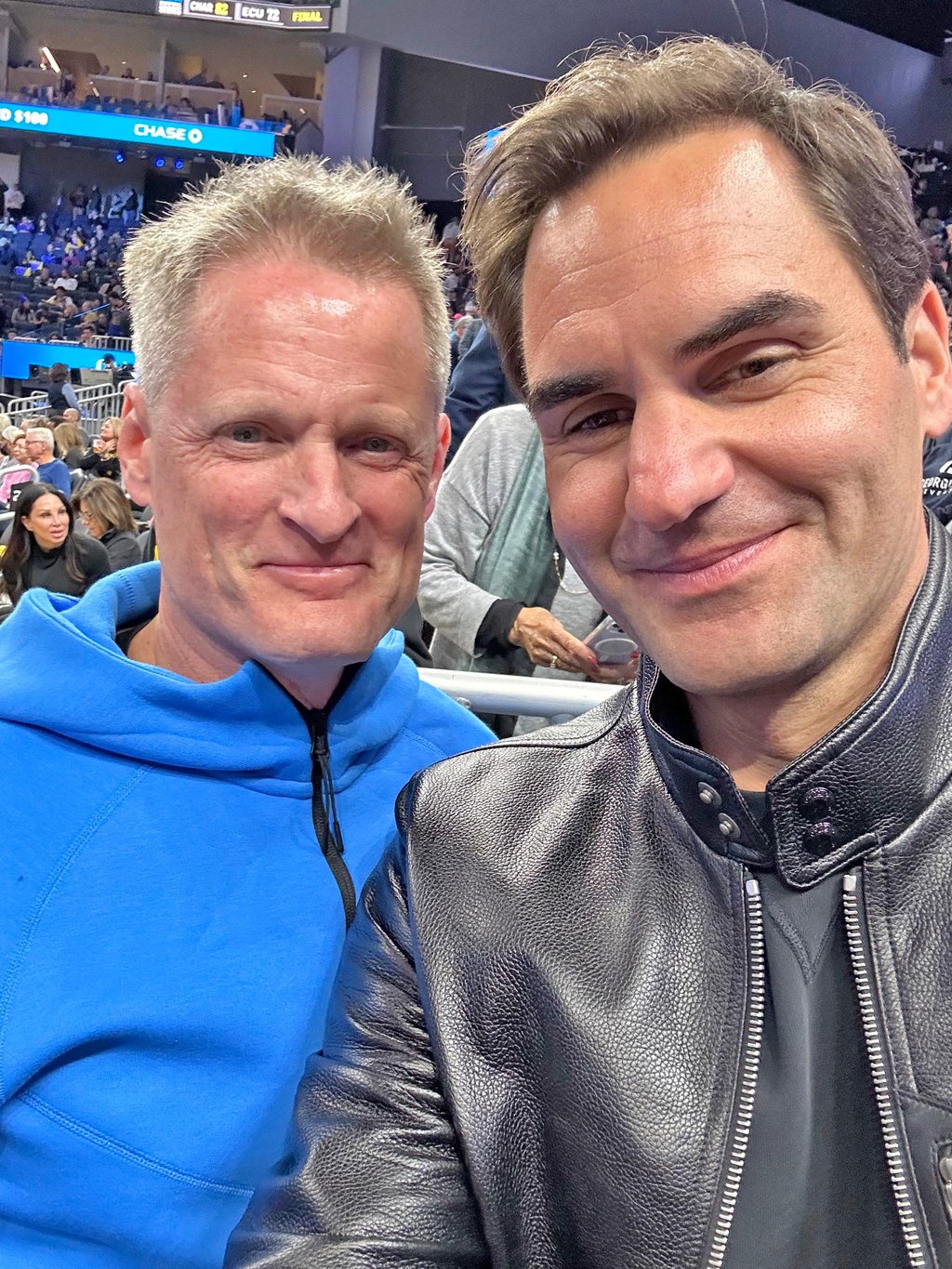 Roger Federer poses for a selfie with a Steve Kerr lookalike at a basketball game.