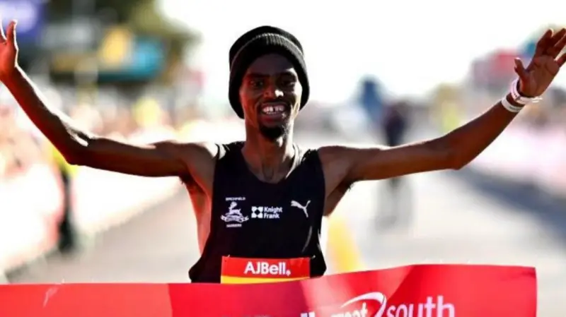 Kadar Omar, wearing a black Puma vest, holding his arms up in celebration as he crosses the finish line. He is wearing a black beanie hat.