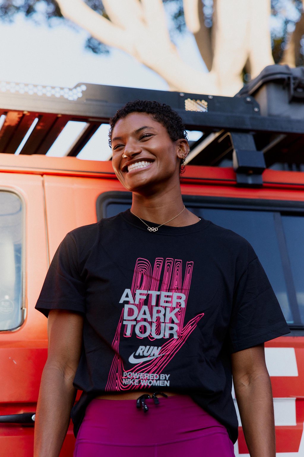 A smiling woman wearing a black t-shirt with pink and silver lettering that reads "After Dark Tour Powered by Nike Women" standing in front of a red vehicle.