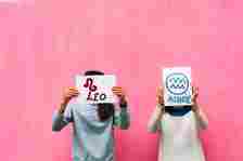 Two individuals standing against a plain background holding signs with zodiac symbols and names: Leo and Aquarius, obscuring their faces