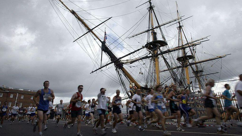 Runners pass by the HMS Victory masted ship in Portsmouth Historic Dockyard. The sky overhead is full of grey clouds.
