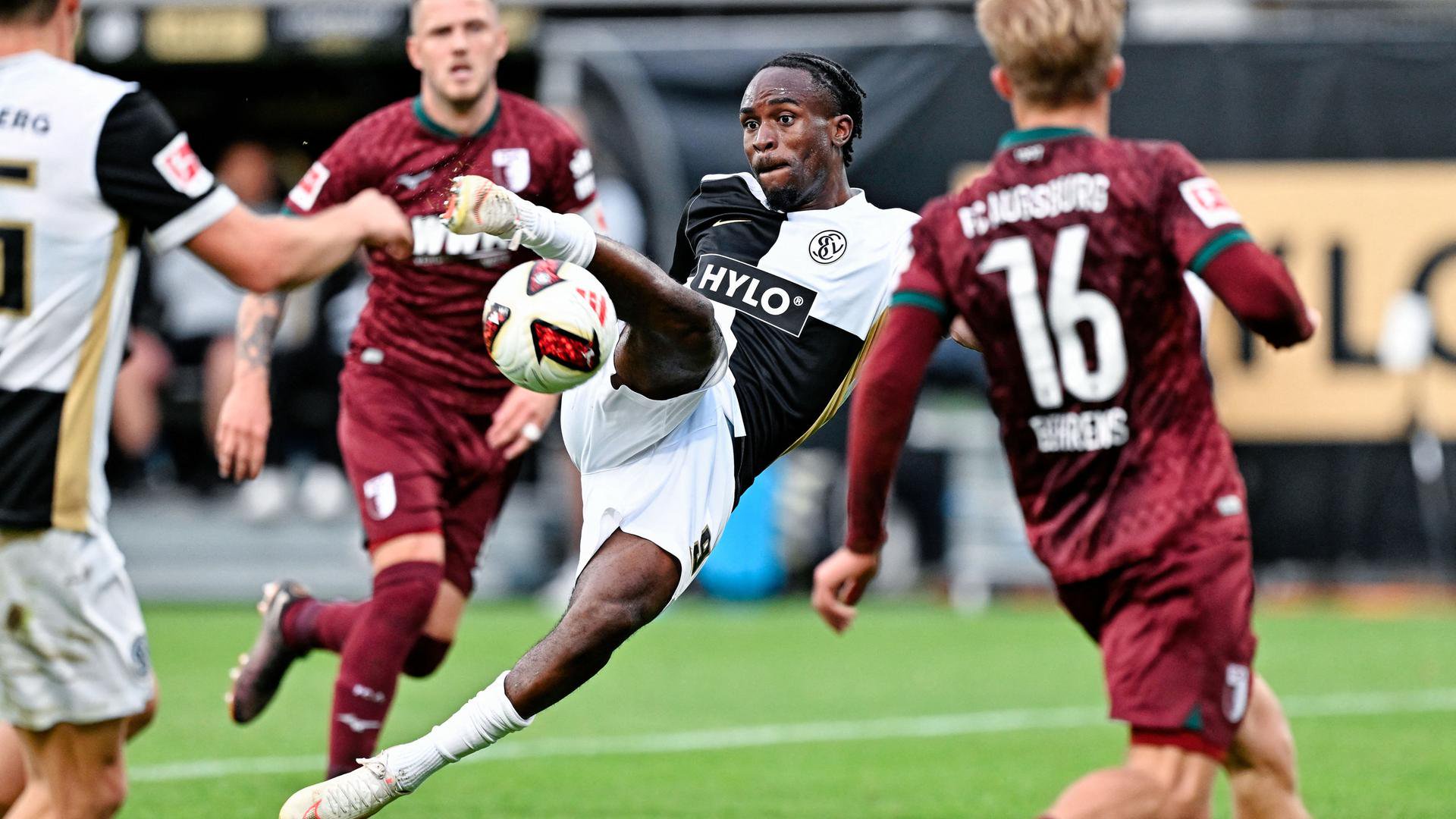 Das Bild zeigt einen Fußball-Spieler, der aus akrobatischer Schräglage heraus zum Torschuss ansetzt, während er von gegnerischen Spielern umringt wird.
