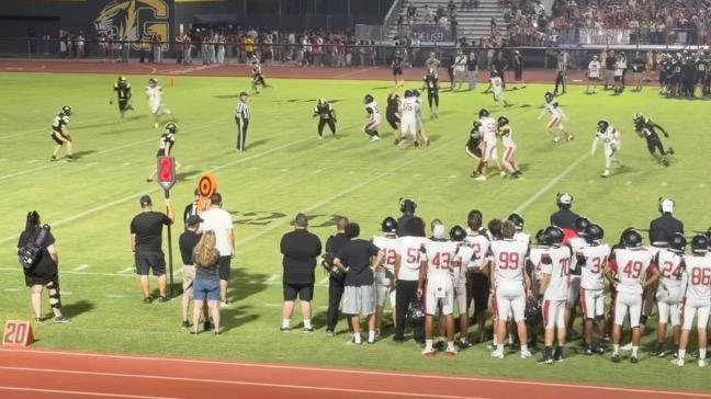 High school football game in progress under the lights, showing players on the field and coaches and players on the sideline.