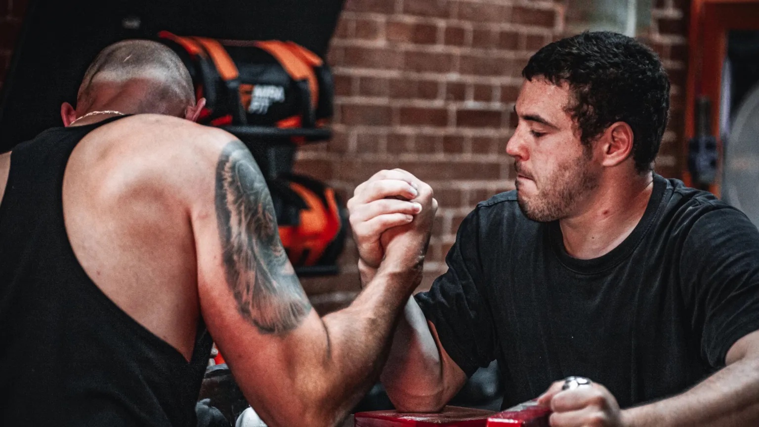 Suffolk Arm Wrestling Team Two men stand with a small table between them arm wrestling. The man furthest away from the camera has dark hair and is wearing a black T-shirt. The man closest to the camera has his face turned away. There is a tiger tattoo on his bicep and he is wearing a black vest top. 