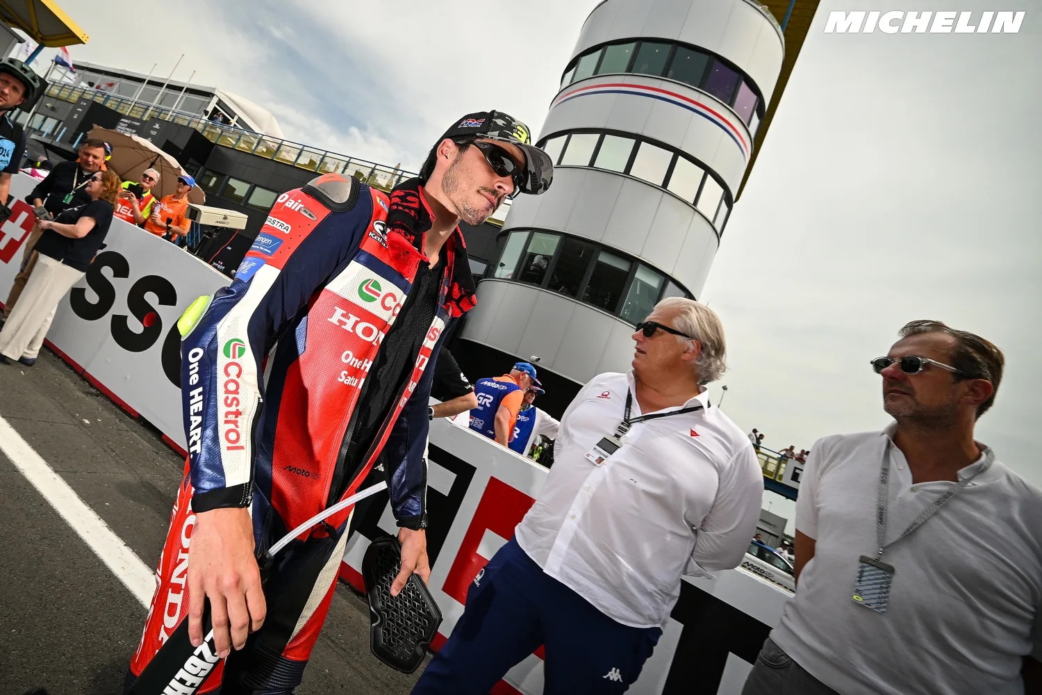 A rider in red, white and blue Honda leathers with Castrol logos walks in the MotoGP paddock while officials stand nearby