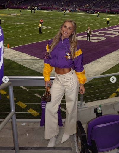A young woman standing in a stadium wearing a purple and yellow jacket, white pants, and holding a small handbag, with the football field in the background.