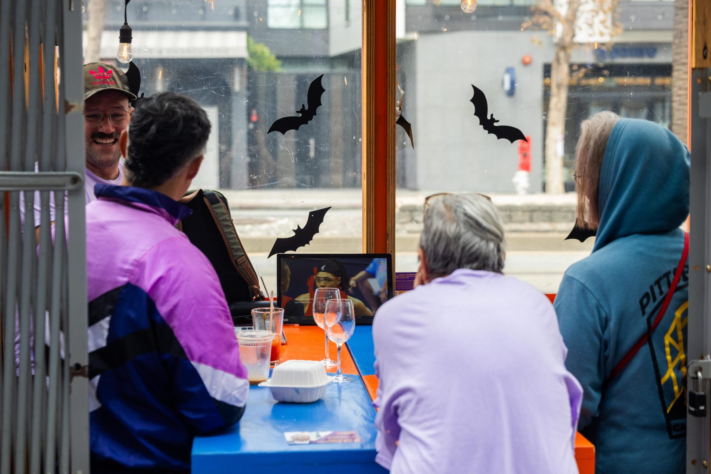 Four people sit at a table in a windowed booth watching a small screen displaying a baseball game.