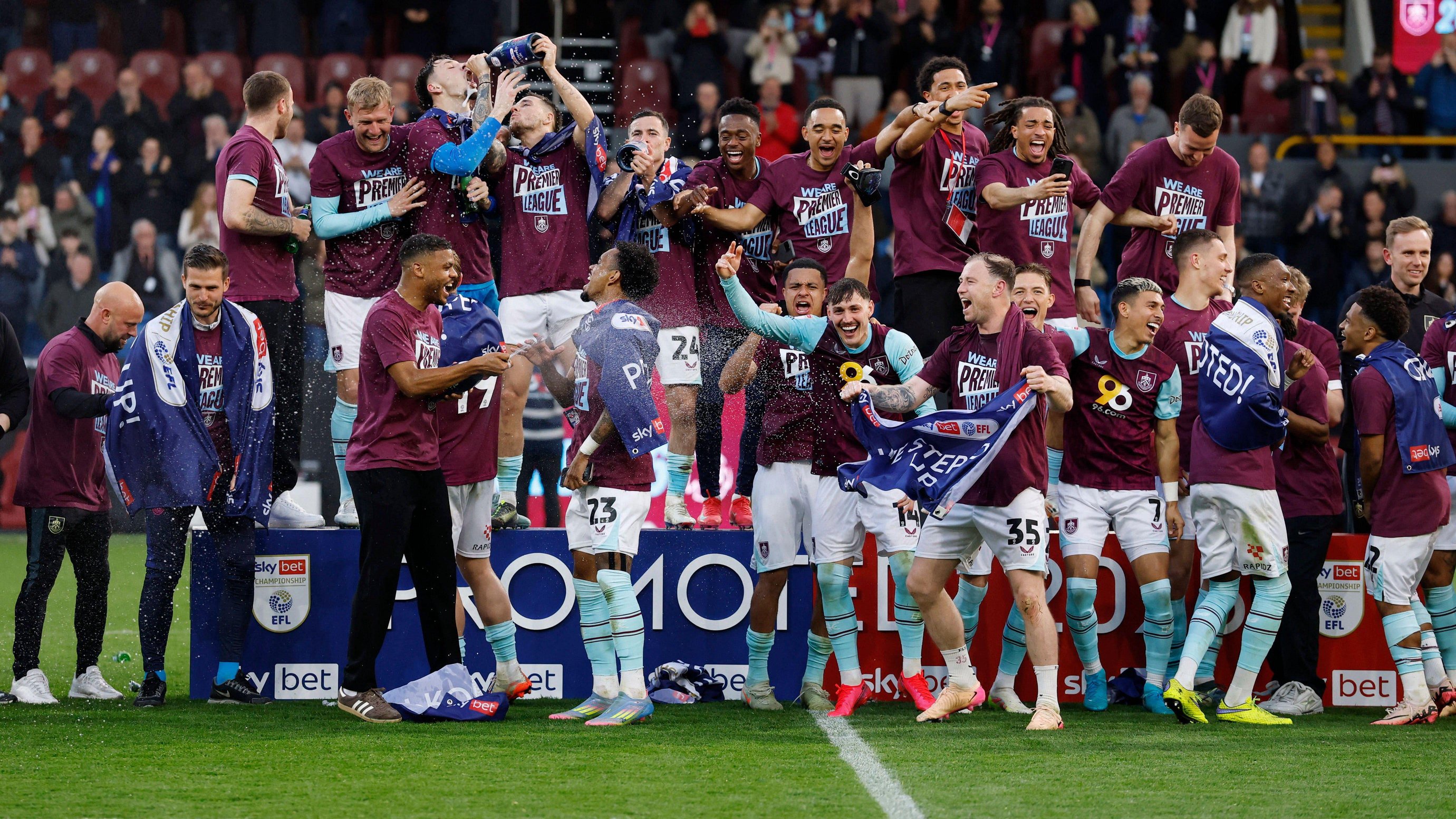 Burnley players celebrate winning promotion to the Premier League in 2025 by swigging from bottles of champagne and wearing claret t-shirts with Premier League written on the them