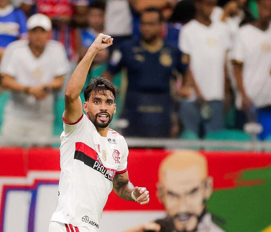 Lucas Paquetá celebra, braço erguido, durante jogo do Flamengo com torcida ao fundo.