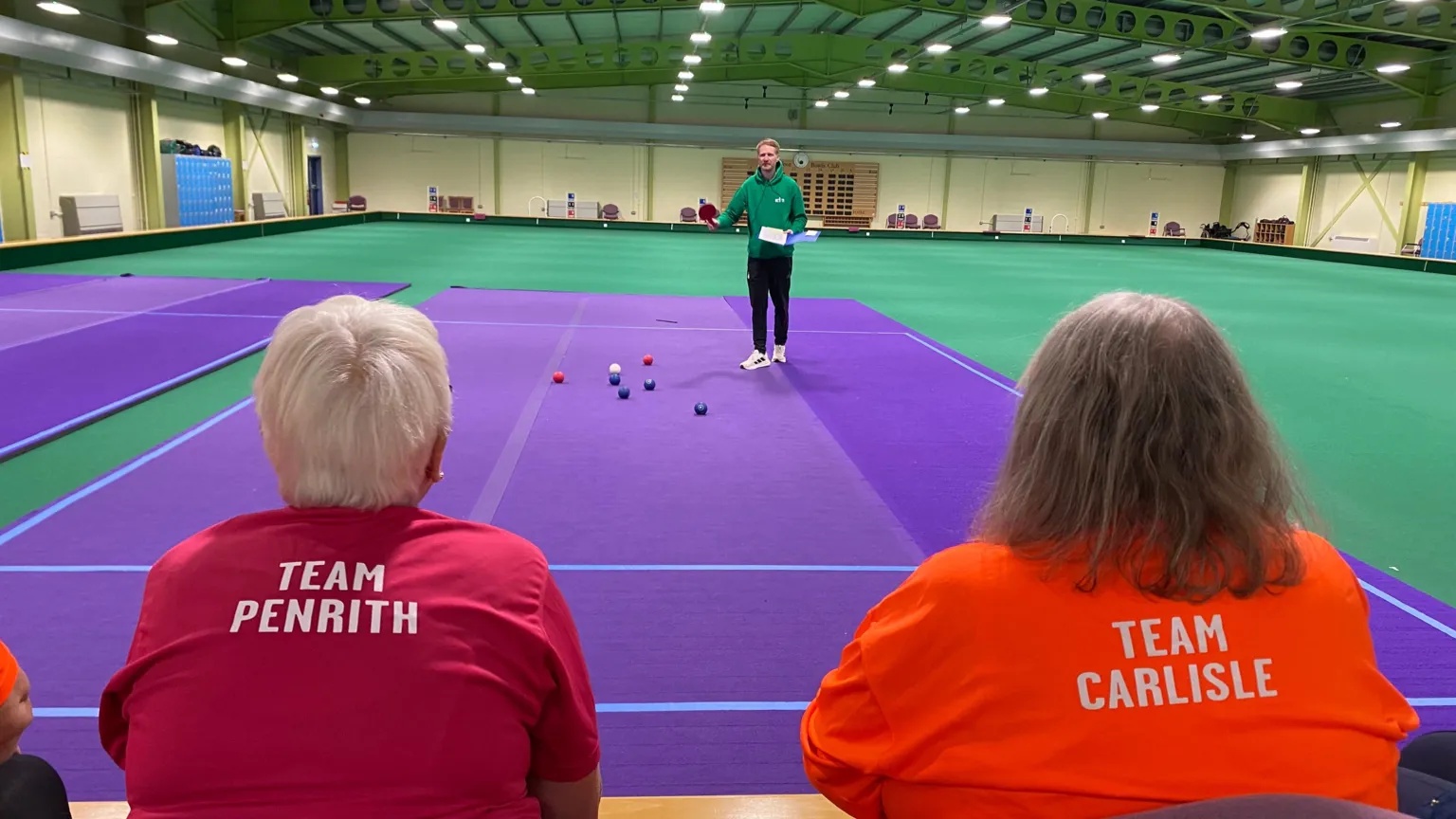 BBC Two competitiors wearing red and orange shirts labelled “Team Penrith" (l) and “Team Carlisle" (r) sit at the edge of a bowls rink. They are facing away from the camera and watching a person standing on a purple playing mat with several bowls in front of them. The large sports hall has green flooring and bright lights overhead.