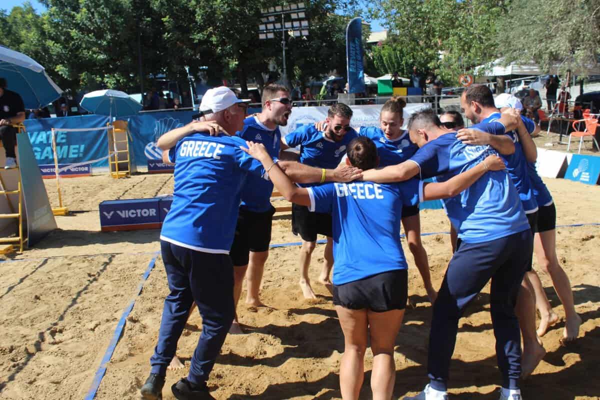 Greek players and coaches celebrating in a circle on the sand court