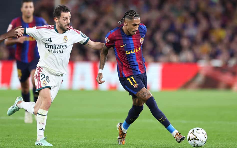 Jogador do Barcelona em uniforme azul e grená dribla a bola enquanto jogador do Real Madrid acompanha a jogada.