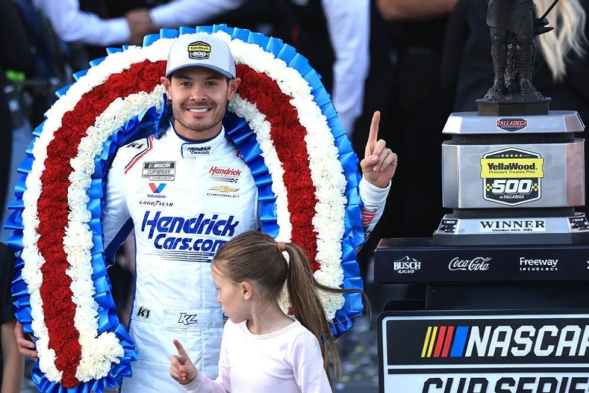 Driver in white Hendrick Motorsports suit with a "Hendrick Cars.com" logo and #5 patch, pointing up beside a wreath and the Talladega winner's trophy at NASC…