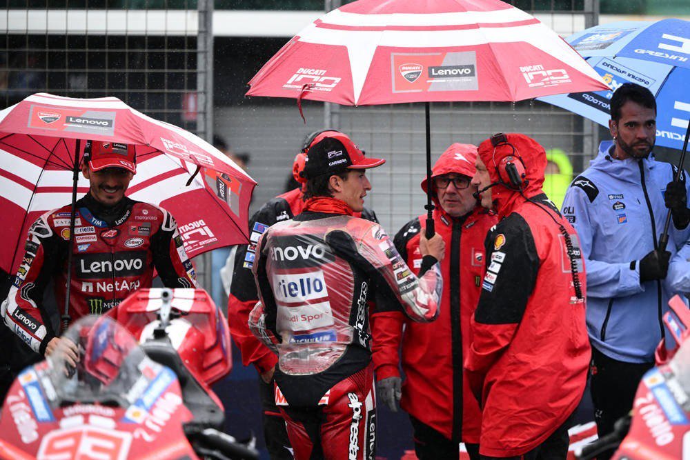 Ducati rider in red and black leathers with a Lenovo and NetApp livery, under a red umbrella on a rainy MotoGP grid, talking with team staff in red rain gear.