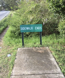 A sidewalk ending abruptly with a green sign that reads &quot;SIDEWALK ENDS&quot; in a grassy area beside a road