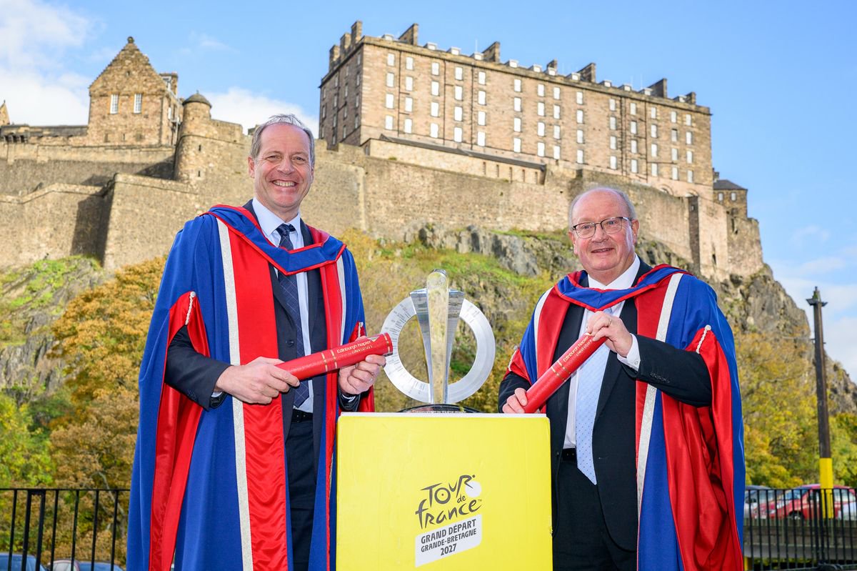 An image of two men wearing red and blue graduation gowns, standing with a trophy in front of a castle.