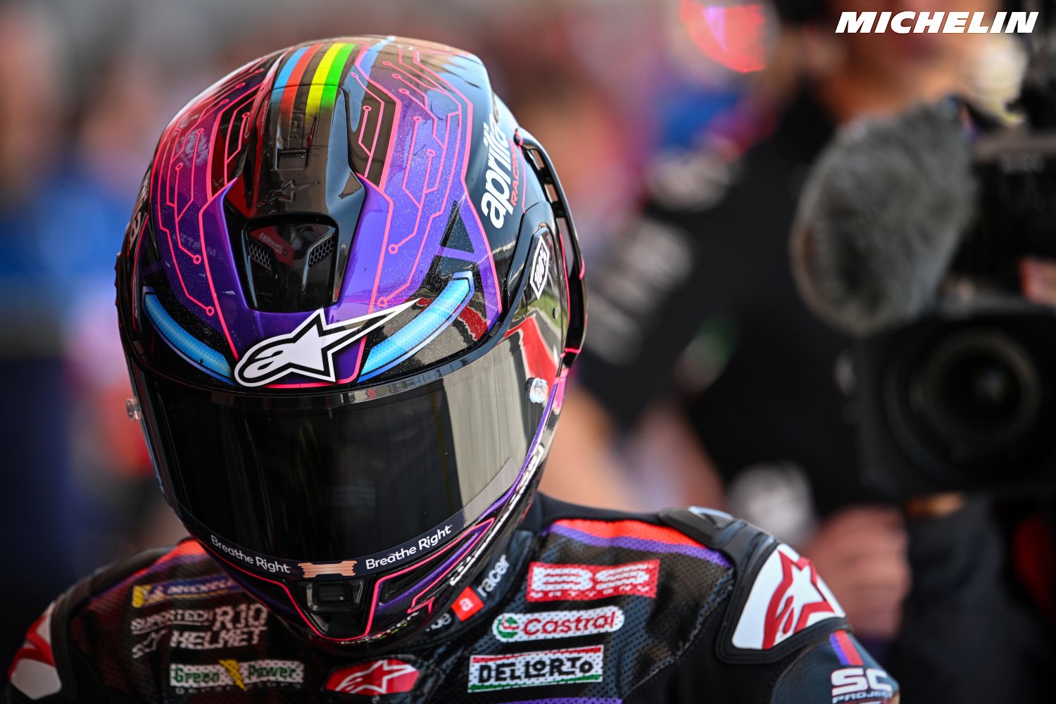 Rider in a purple-and-pink Aprilia-liveried helmet with Alpinestars branding, speaking near a TV camera in the MotoGP paddock