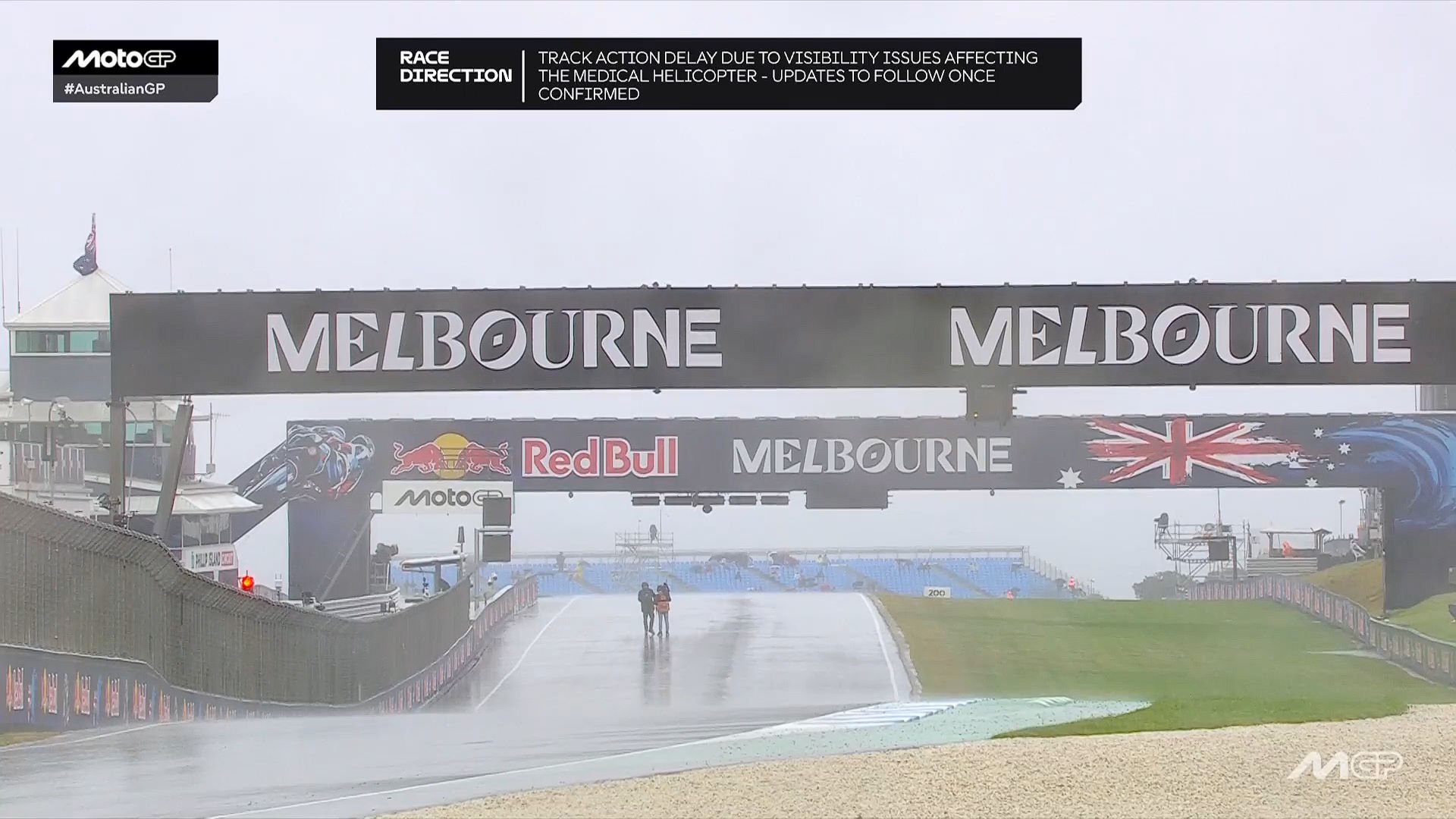 Two people in rain gear walk across a wet MotoGP track at Phillip Island, Melbourne, under a grey sky during the weather delay.