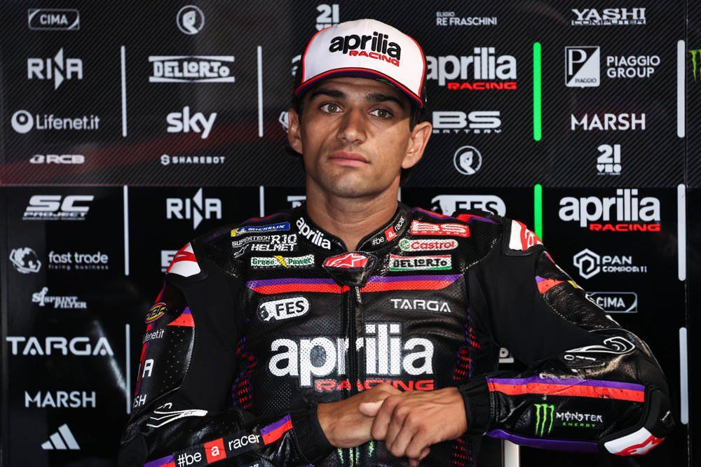 A rider in Aprilia Racing leathers and cap stands in the pit garage, arms resting on a wall of team sponsor logos, in the MotoGP paddock.