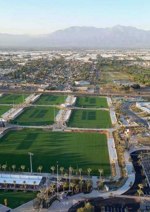 Aerial view of the Ontario Sports Empire facility featuring multiple green fields with long shadows, surrounded by palm trees, parking areas, and a city view with mountains in the background.