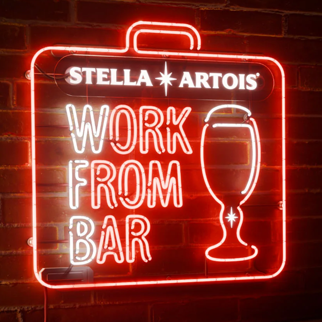 Neon sign "Work from Bar" with Stella Artois bottle and glass near laptop on bar counter.