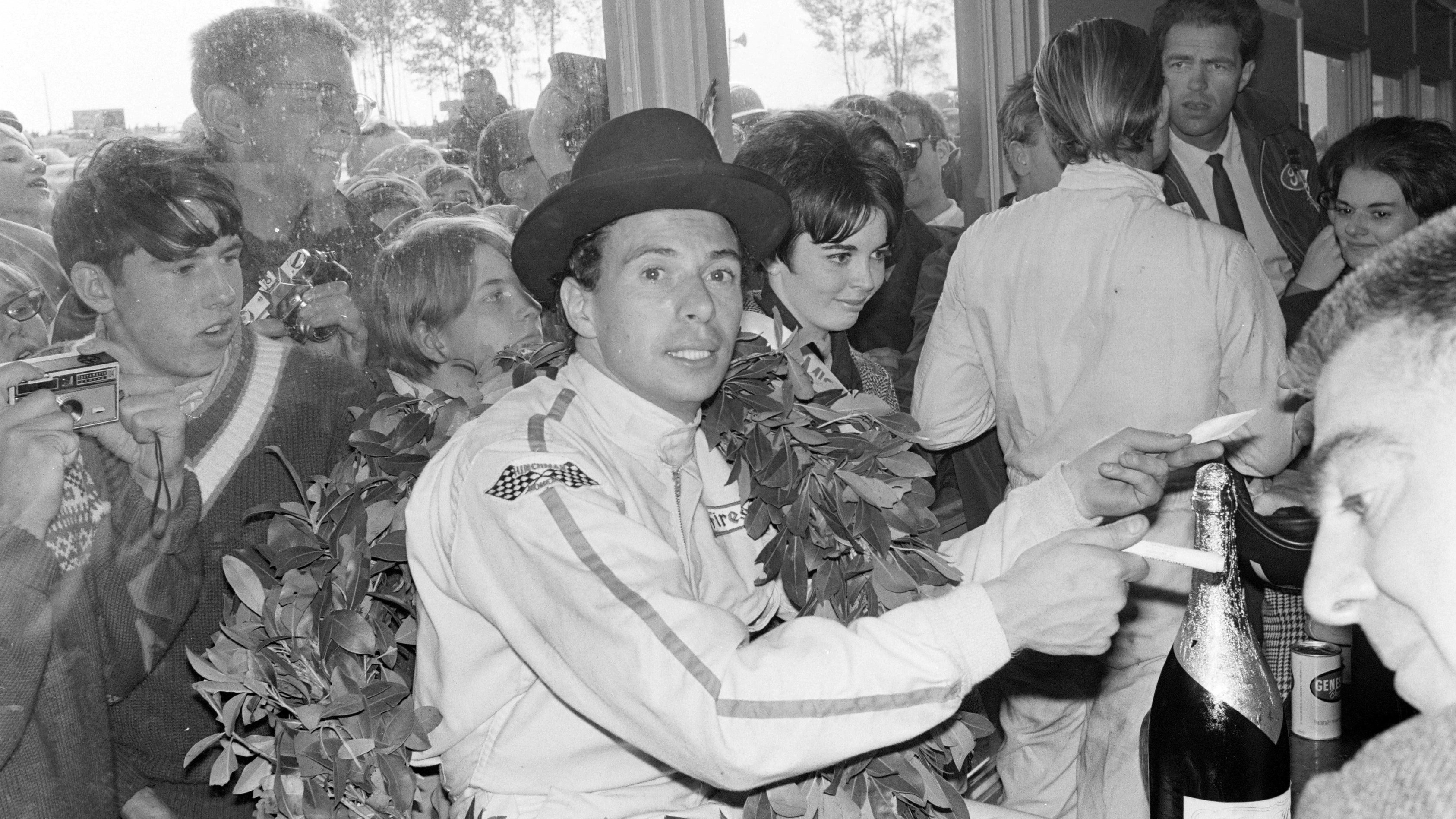 Jim Clark, wearing a hat, celebrates victory at Watkins Glen after the podium celebration in 1967. Fans are pressed against a window outside, trying to get a glimpse of him