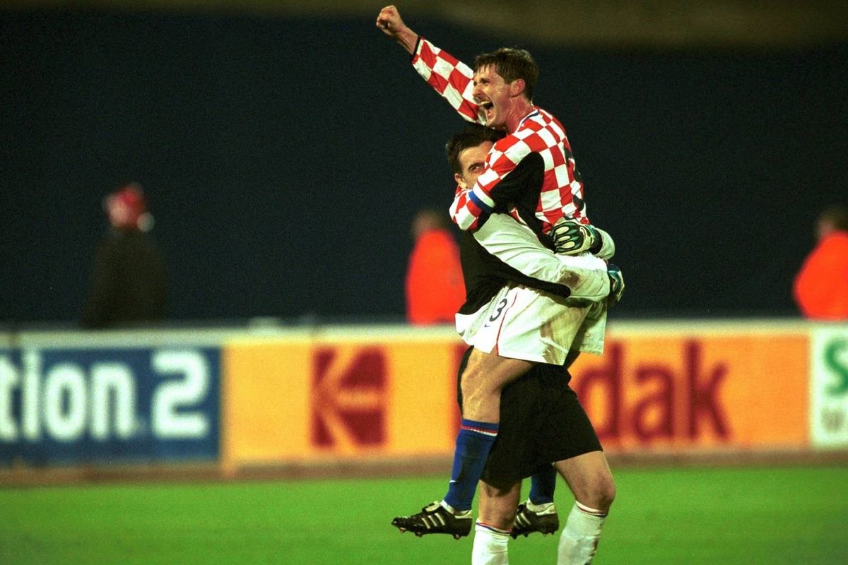 Two soccer players are celebrating on a field, one player is airborne and the other is holding him aloft. The background features a blurred advertisement banner with the word "dak" prominently displayed.