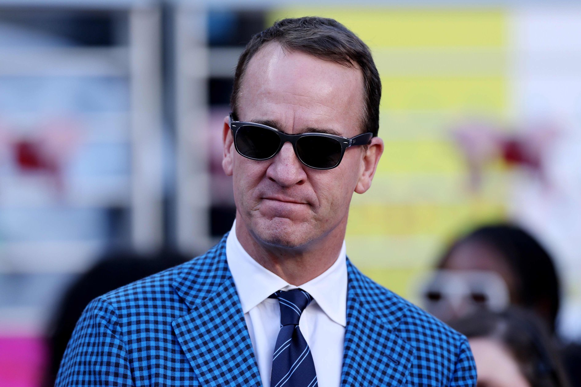Peyton Manning wears sunglasses and a blue gingham suit jacket.